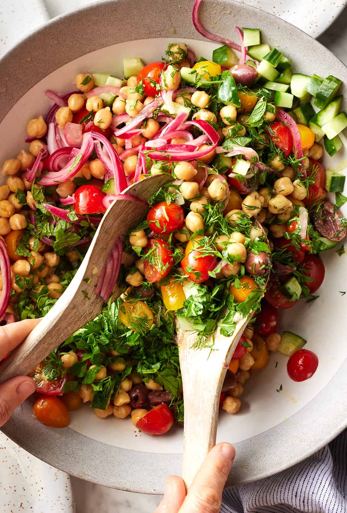 Hands using wooden spoons to toss Mediterranean chickpea salad in large bowl